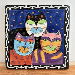May include: A square ceramic decorative plate featuring three stylized cat portraits. The cats have colorful faces and are set against a dark blue background with yellow dots. The plate has a black border with white polka dots.