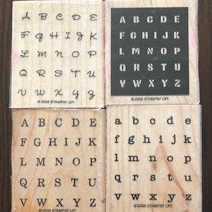 May include: Four wooden alphabet stamps. Two stamps feature uppercase letters in a serif font, one in a stencil font, and one in a cursive font. The lower right stamp has lowercase letters. Each stamp has the text "©2005 STAMPIN' UP!"