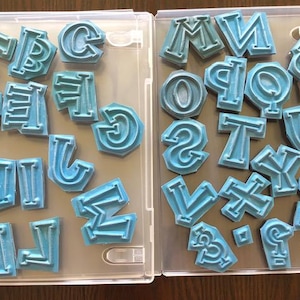 May include: A set of blue alphabet letter stamps in two clear plastic storage cases. The letters are in uppercase and are arranged alphabetically in each case.