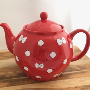 May include: Red ceramic teapot with white polka dots and bows. The teapot has a lid and a handle.