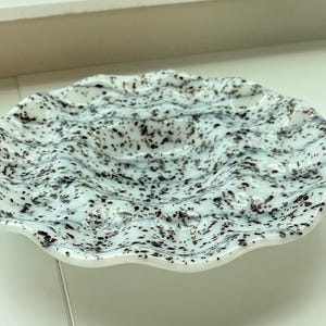 May include: A decorative, speckled glass plate with a wavy rim. The plate features a light blue base with black speckles, creating a unique pattern. The plate's design is reminiscent of ocean waves.