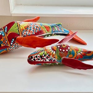 May include: Two decorative ceramic whale figurines. The whales are painted with vibrant colors, including red, blue, green, and yellow. The fins and tails are a bright red. The whales are resting on a white surface.