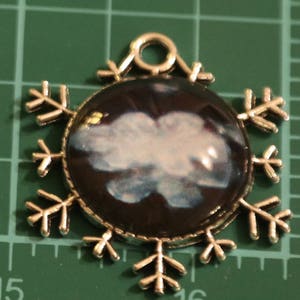 May include: A snowflake pendant with a gold-colored frame and a dark background. The center features a white and blue cloud design. The pendant has a loop at the top for hanging.