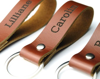 May include: Three brown leather keychains with silver key rings. Each keychain has a name engraved on it: Lillian, Carolin, and Brad. The keychains are made of a smooth, light brown leather.