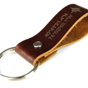 Könnte beinhalten: Brauner Leder-Schlüsselanhänger mit einem silbernen Ring und einem Kompass-Symbol. Der Schlüsselanhänger ist mit den Koordinaten "40.4121.47N 74.0240.17W" graviert.