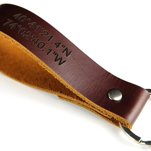 Könnte beinhalten: Ein brauner Leder-Schlüsselanhänger mit einem silbernen Schlüsselring. Das Leder ist mit den Koordinaten "40.4121.4'N 74.0240.1'W" graviert.