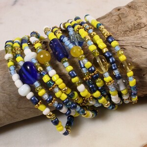 May include: A set of beaded bracelets in yellow, blue, and white. The bracelets are stacked on top of each other and feature small glass beads.