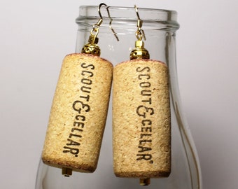May include: A pair of gold-toned earrings with cork dangles. The cork is engraved with the text "Scout & Cellar."