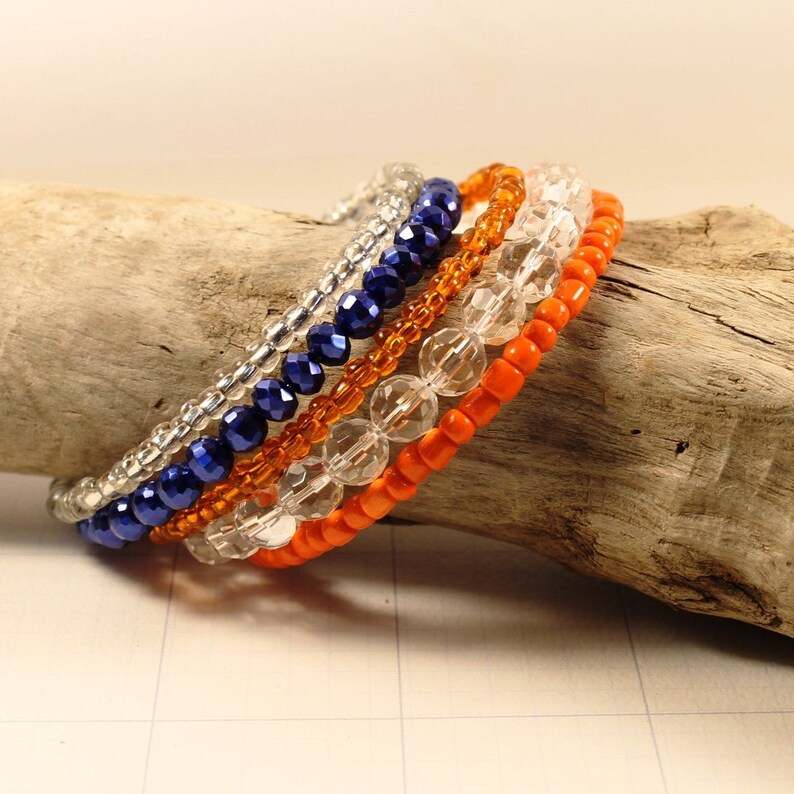 May include: Five beaded bracelets stacked on a piece of driftwood. The bracelets are made of silver, orange, and blue beads.