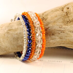May include: Set of five beaded bracelets in orange, clear, and blue. The bracelets are stacked on a piece of driftwood.