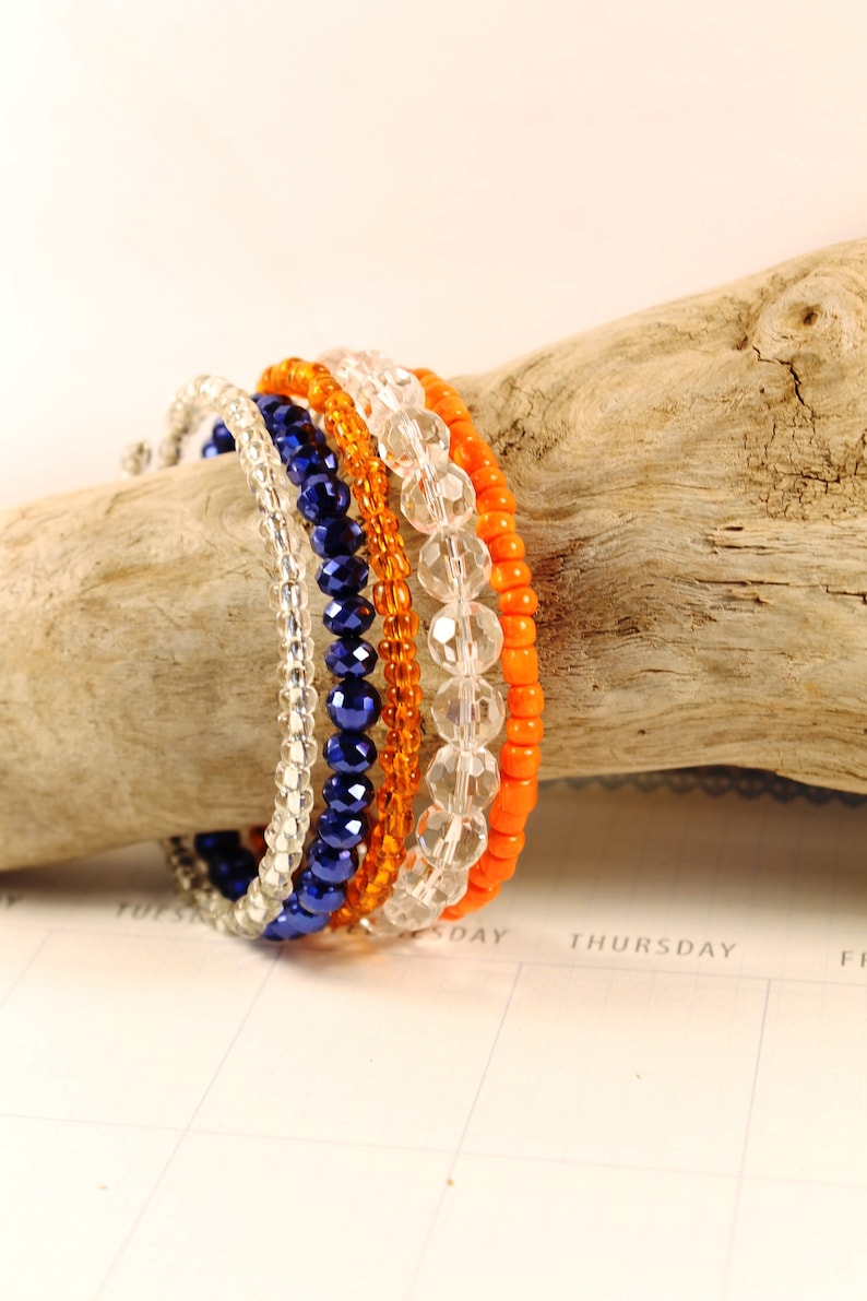 May include: Set of five beaded bracelets in orange, clear, and blue. The bracelets are stacked on a piece of driftwood.