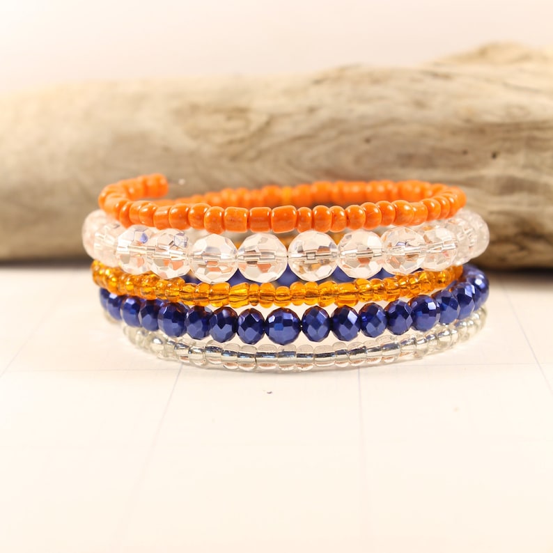 May include: A set of six beaded bracelets in orange, clear, gold, blue, and silver. The bracelets are stacked on top of each other.