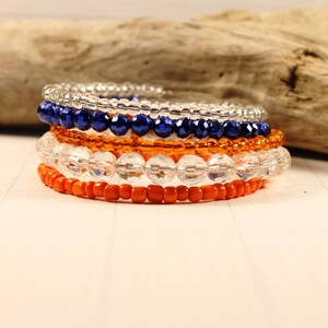 May include: A set of five beaded bracelets in orange, white, and blue. The bracelets are stacked on top of each other, creating a layered look.