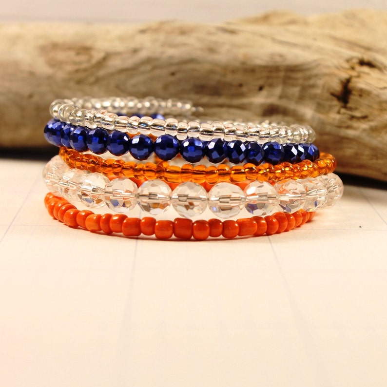 May include: A set of five beaded bracelets in orange, white, and blue. The bracelets are stacked on top of each other, creating a layered look.