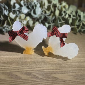May include: Two decorative rooster figurines. The roosters are white with a textured surface, featuring a red and black plaid bow. The base and feet are a golden color. The figurines are displayed on a wooden surface.