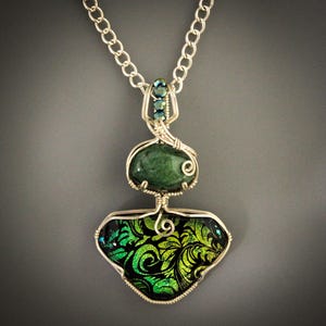 May include: A silver wire-wrapped pendant necklace featuring a smooth, oval-shaped dark green stone positioned above a larger, triangular-shaped black glass piece with a vibrant green and yellow swirling floral pattern, suspended on a silver chain.