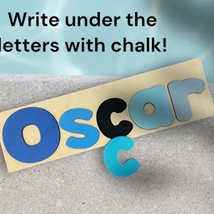 May include: A wooden board with the name "Oscar" spelled out in blue, black, and light blue letters. The letters are raised and have a textured surface. The text "Write under the letters with chalk!" is written above the name.