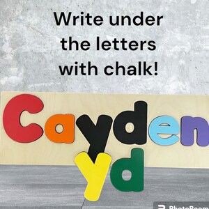 May include: Wooden puzzle with the name Cayden spelled out in colorful letters. The letters are cut out and can be removed. The letters are red, orange, black, light blue, and purple. The background is light wood. The text "Write under the letters with chalk!" is written above the puzzle.
