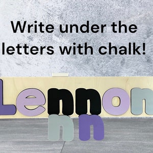 May include: Wooden name puzzle with the name Lennon spelled out in purple, black, and gray letters. The letters are on a light wood background. The text "Write under the letters with chalk!" is above the puzzle.