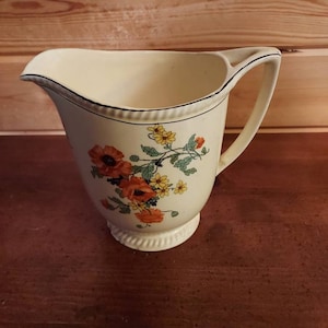 May include: Cream-colored ceramic pitcher with a floral design featuring orange and yellow flowers. The pitcher has a black trim along the rim and handle. The base is slightly raised with a decorative edge. A classic piece for serving.