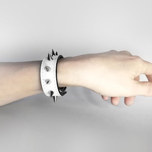 May include: White leather bracelet with black accents and silver spikes. The bracelet is worn on a person's wrist.