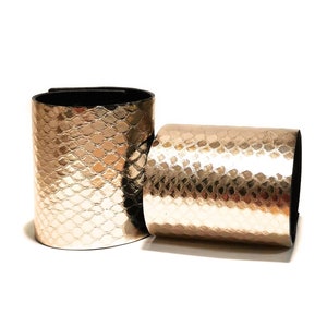 May include: Two gold-colored, textured leather wrist cuffs. One cuff is upright, and the other is lying on its side. The cuffs have a scale-like pattern and black lining. The cuffs are accessories.