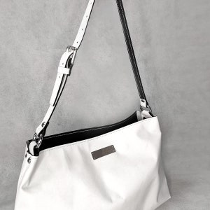 May include: White leather shoulder bag with a black leather trim and a silver buckle. The bag has a small metal tag with the word "Atelier" on it.