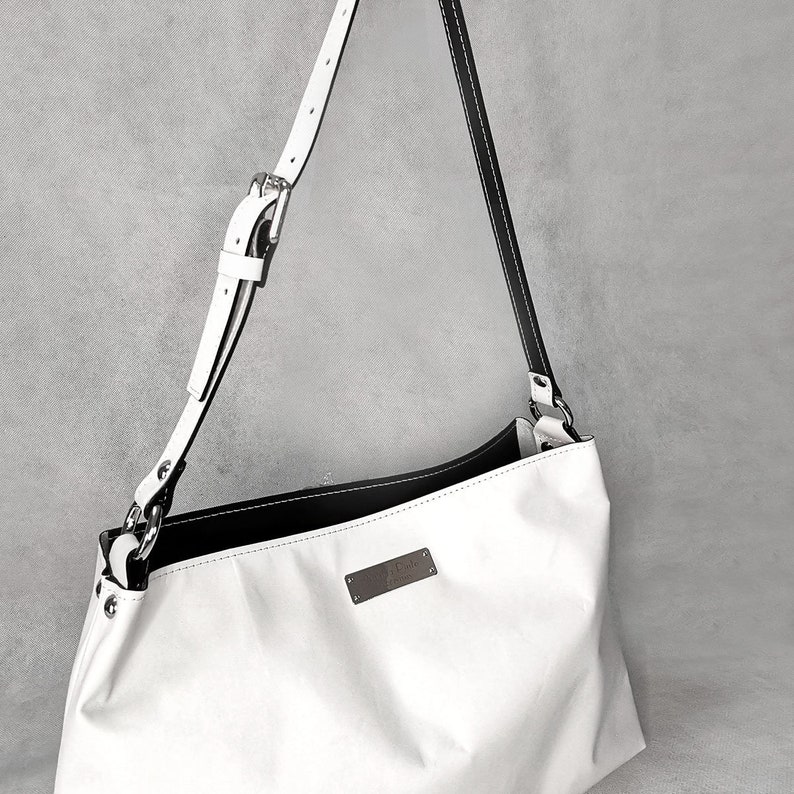 May include: White leather shoulder bag with a black leather trim and a silver buckle. The bag has a small metal tag with the word "Atelier" on it.