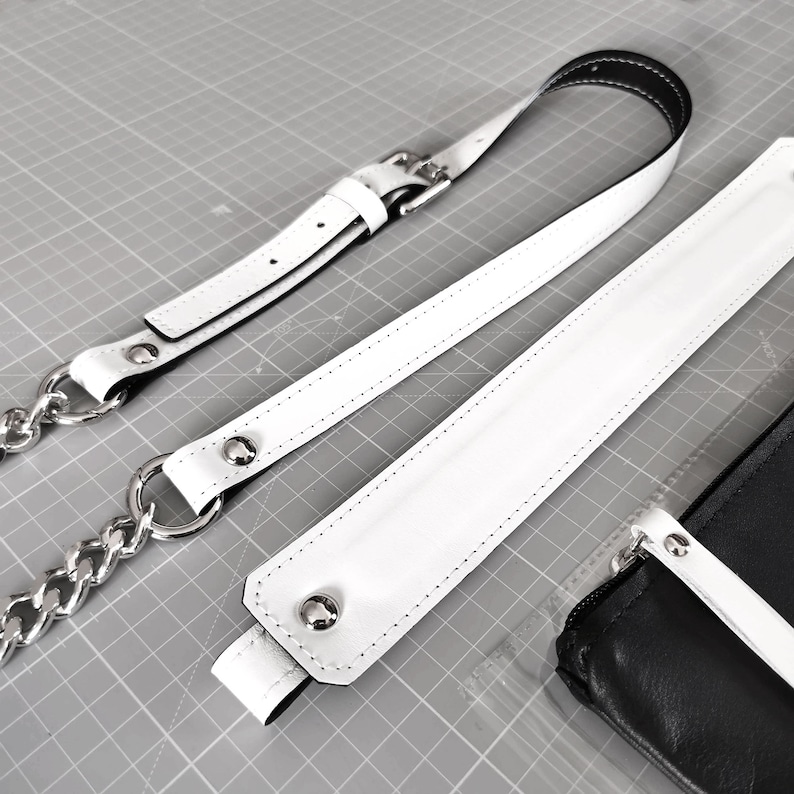 May include: Three white leather straps with silver metal hardware. The straps have stitching along the edges. The straps are on a gray surface.