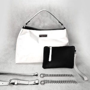 May include: A white leather tote bag with a black leather pocket and a silver chain strap. The bag has a silver logo that says "Angela Davis" on the front.