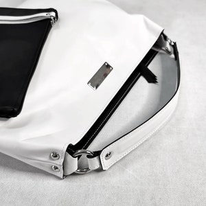May include: White leather handbag with a black zipper and a black strap. The bag has a silver metal plate with the brand name on it.