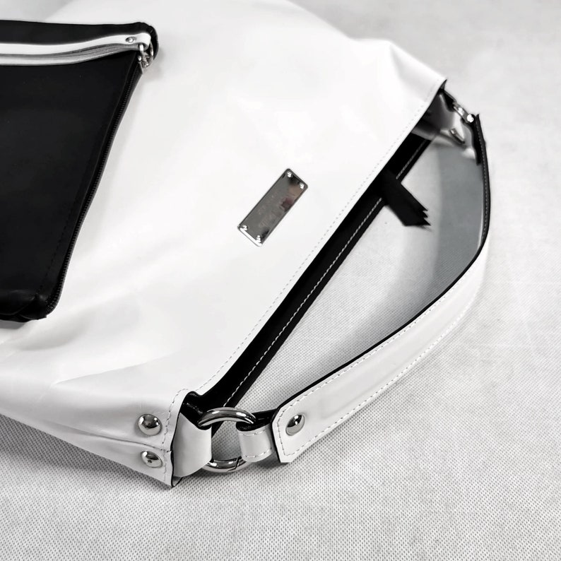 May include: White leather handbag with a black zipper and a black strap. The bag has a silver metal plate with the brand name on it.