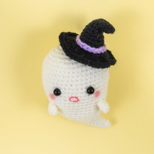 Puede incluir: Un fantasma blanco de crochet que lleva un sombrero de bruja negro con una banda morada. El fantasma tiene una cara linda con ojos negros y una boca rosa.