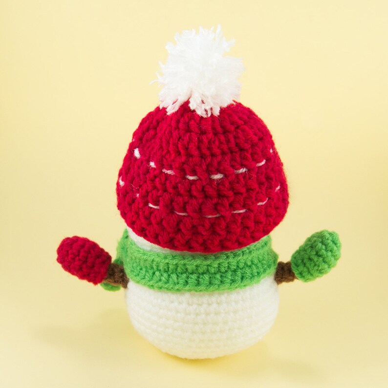 May include: A crocheted snowman wearing a red and white hat with a white pom-pom and a green scarf. The snowman has two small crocheted arms.