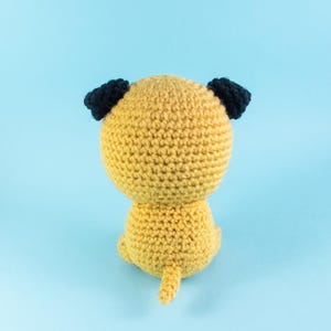 May include: A crocheted stuffed dog toy with a yellow body and black ears. The dog is sitting on a light blue background.