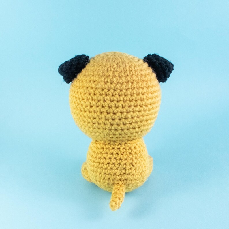 May include: A crocheted stuffed dog toy with a yellow body and black ears. The dog is sitting on a light blue background.