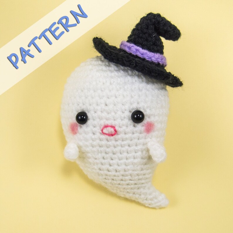 Puede incluir: Un fantasma blanco de crochet que lleva un sombrero de bruja negro con una banda morada. El fantasma tiene ojos negros, mejillas rosadas y una boca roja.