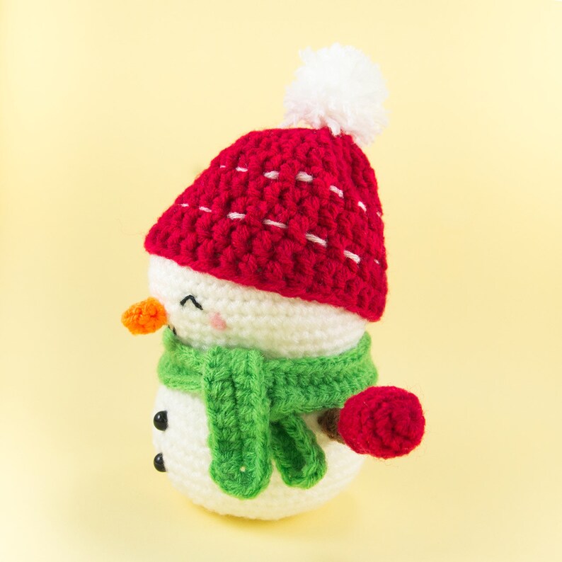 May include: A crocheted snowman wearing a red hat with white pom pom and a green scarf. The snowman has a carrot nose and black eyes.