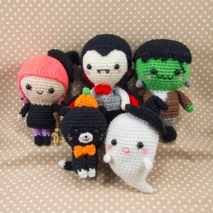 May include: Five crocheted Halloween characters: a vampire, a ghost, a witch, a black cat, and a Frankenstein. The characters are all smiling and have big eyes.