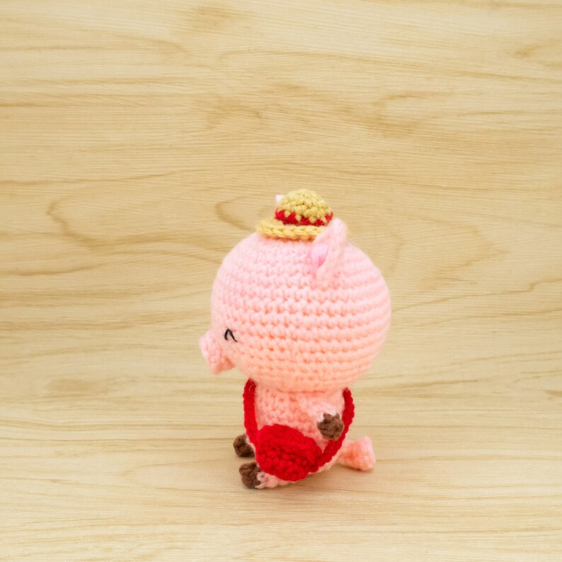 May include: A pink crocheted pig wearing a straw hat with a red and yellow band. The pig is holding a red crocheted bag.