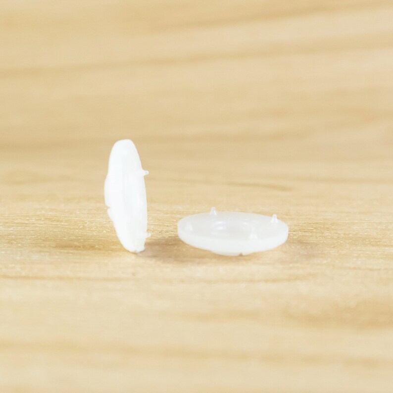 May include: Two white plastic snap fasteners, one lying flat and the other standing upright on a wooden surface.