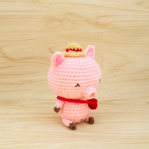 May include: A pink crocheted pig wearing a straw hat and a red scarf. The pig is sitting on a light brown wooden surface.