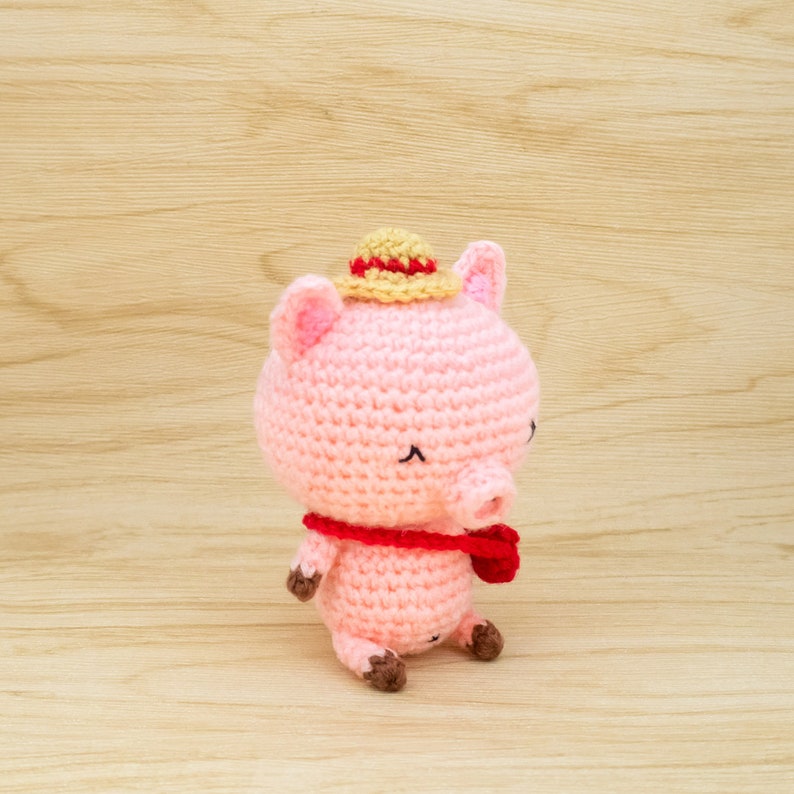 May include: A pink crocheted pig wearing a straw hat and a red scarf. The pig is sitting on a light brown wooden surface.