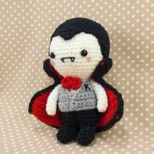 May include: A crocheted vampire doll wearing a red cape and a gray vest with a black letter K. The doll has black hair, a white face, and black eyes.