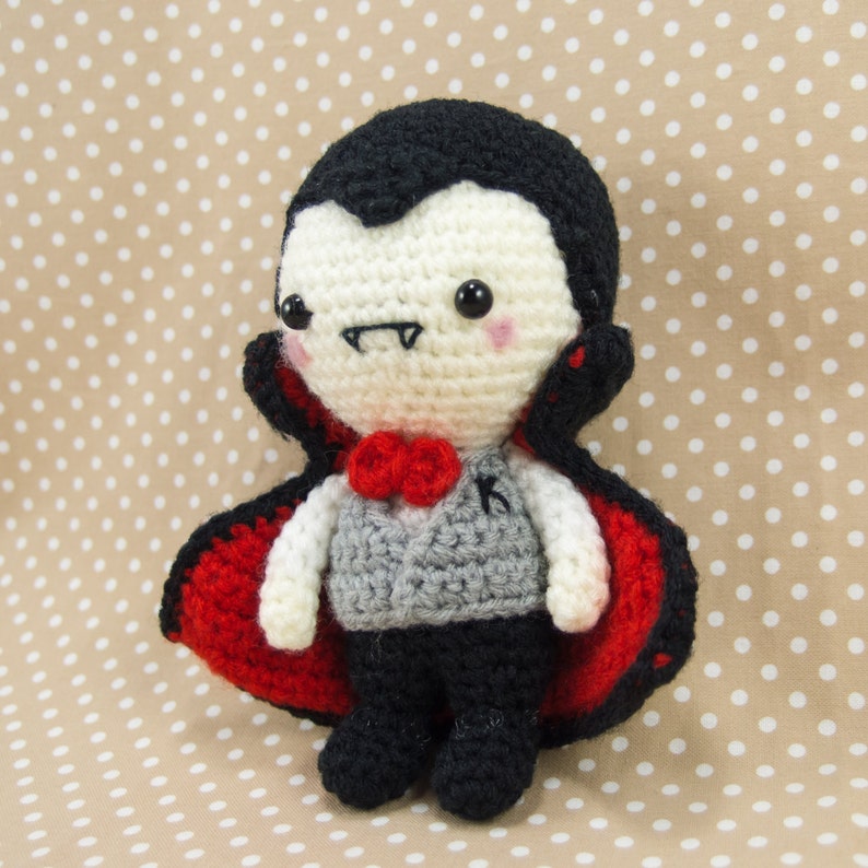 May include: A crocheted vampire doll wearing a red cape and a gray vest with a black letter K. The doll has black hair, a white face, and black eyes.