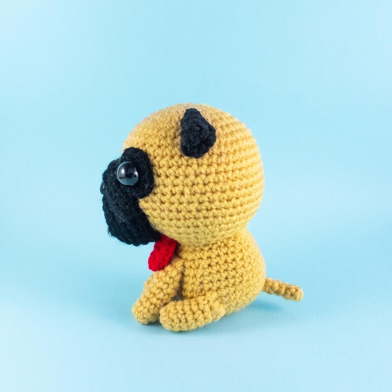 May include: A crocheted pug dog toy with black ears, black eyes, and a red bow tie. The toy is sitting on a light blue background.