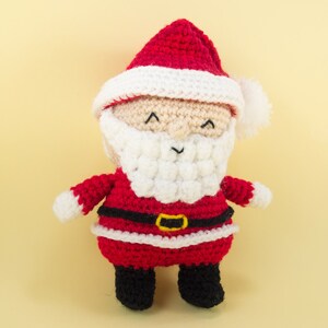 Pode incluir: Um boneco de Papai Noel de croch&ecirc; com um chap&eacute;u vermelho, barba branca e cinto preto. O boneco est&aacute; vestindo um terno vermelho com detalhes brancos.