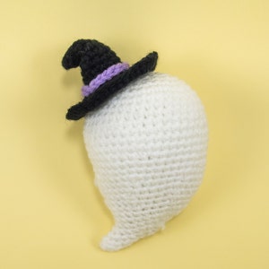 Puede incluir: Un fantasma blanco de crochet que lleva un sombrero de bruja negro con una banda morada. El fantasma tiene una cola larga.