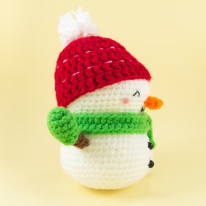 May include: A crocheted snowman wearing a red hat with a white pom-pom and a green scarf. The snowman has a carrot nose and black eyes.