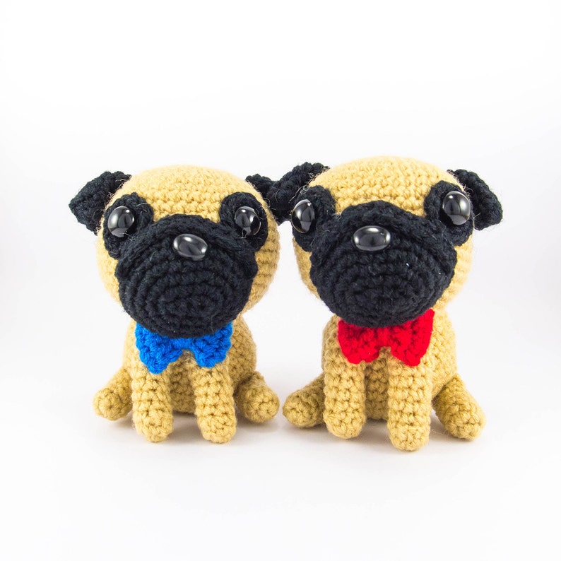 May include: Two crocheted pug dog plushies, one with a blue bow tie and the other with a red bow tie, sitting side by side on a white background.
